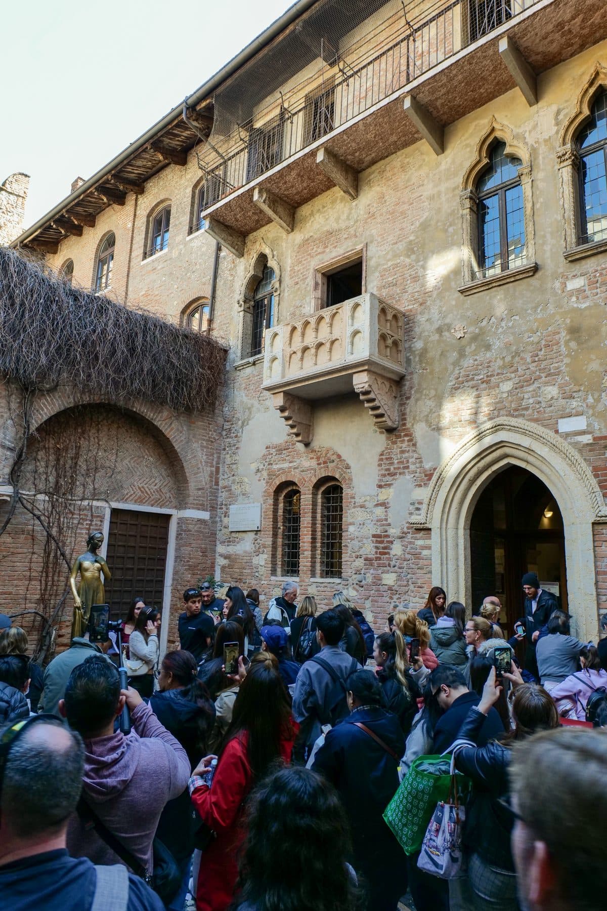 Visitors crowding the courtyard of the house of Juliet in Verona, with its stone balcony above a pointed archway and a bronze statue on the left