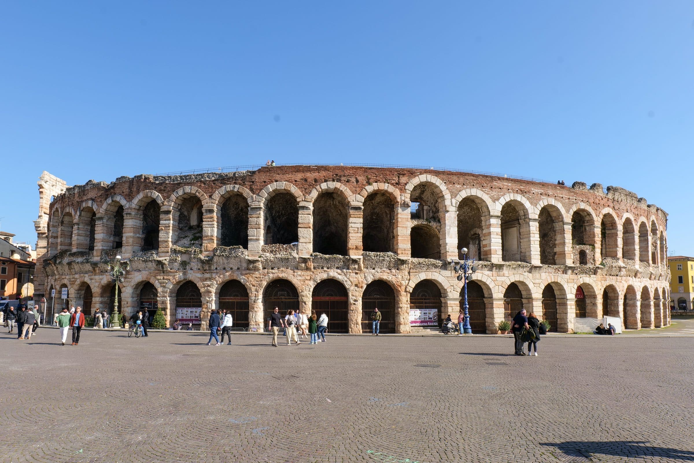 The Roman Arena in Piazza Bra, Verona, under a clear blue sky, with walkers on the cobbled square in front of its two tiers of arches