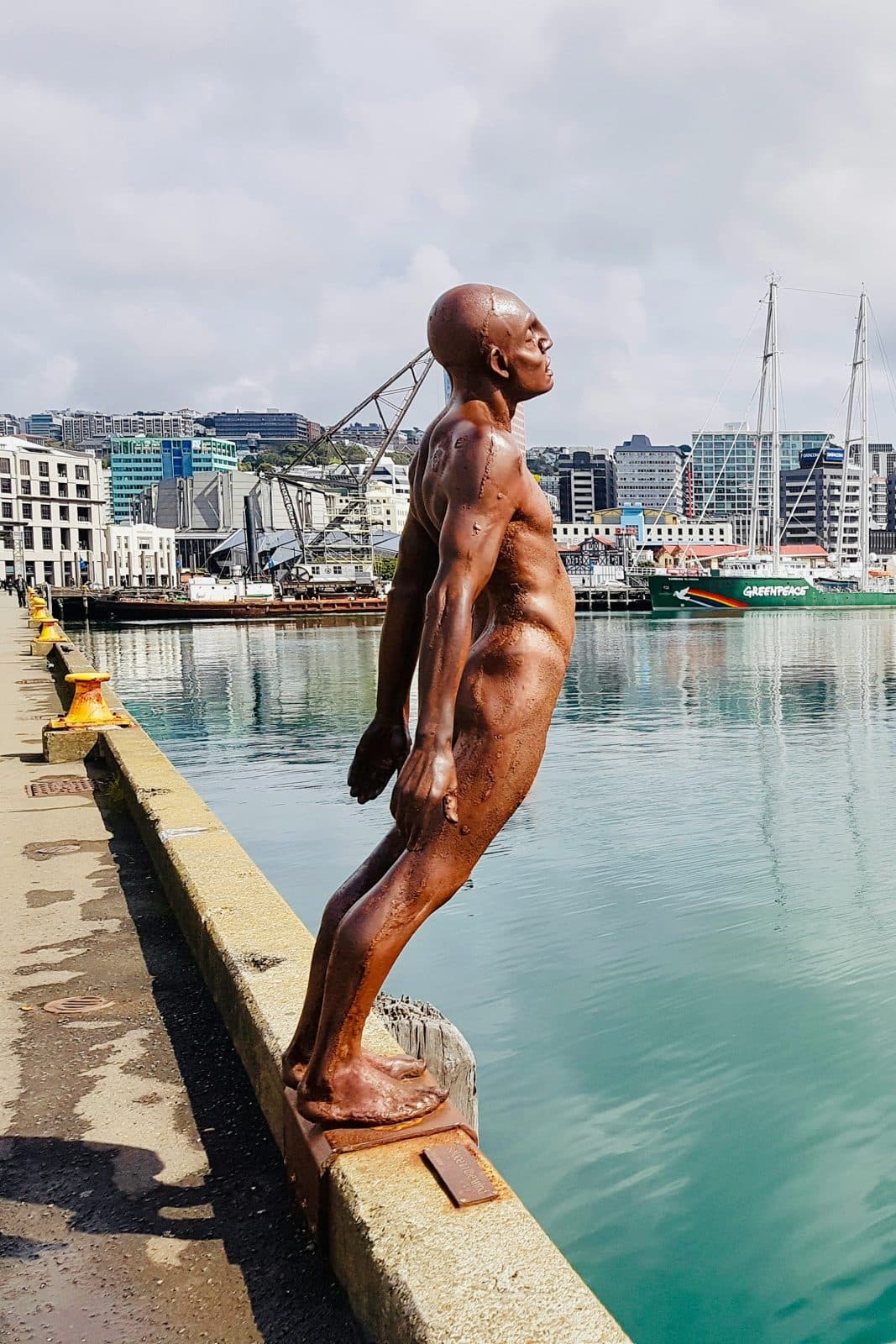 A bronze figure leaning back at the edge of a wharf on Wellington's waterfront, with the harbour and city buildings behind, on the route of the marathon